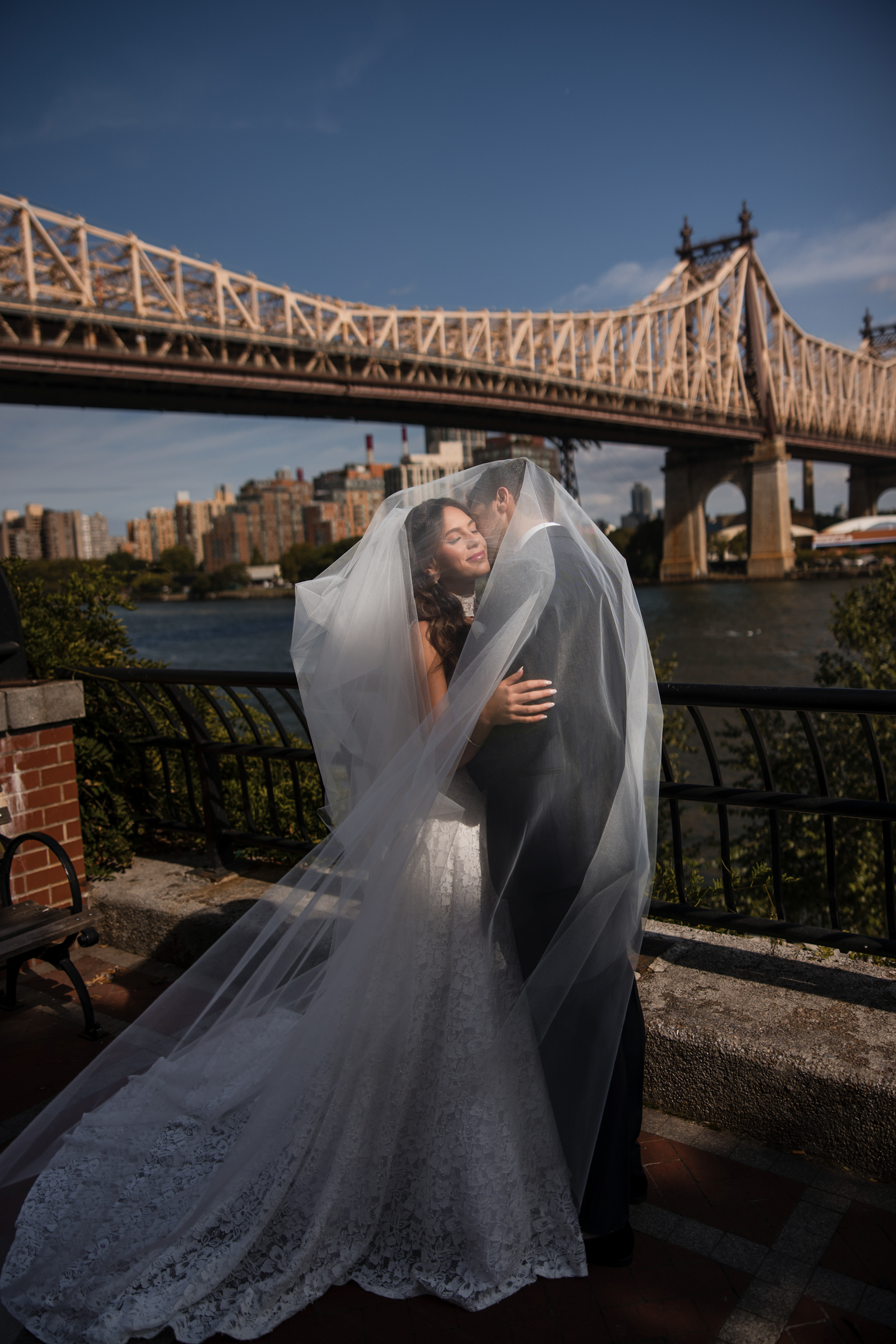 A bride and groom embrace outdoors under a long veil, with a large city bridge and river in the background on a sunny day, capturing the romantic elegance of a Guastavino’s wedding.