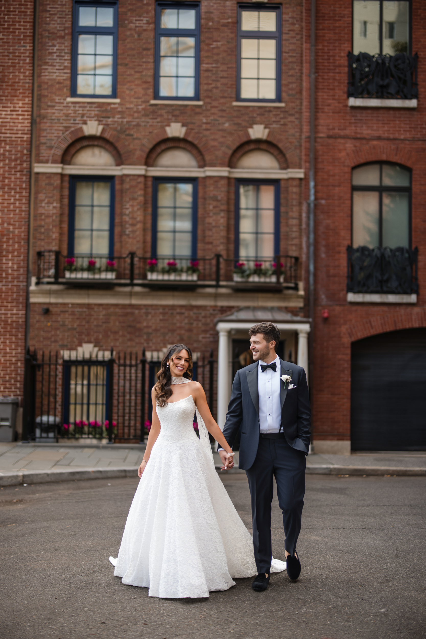 A bride in a white gown and a groom in a tuxedo walk hand in hand on a city street in front of a red brick building, capturing the timeless elegance of a Guastavino’s wedding.