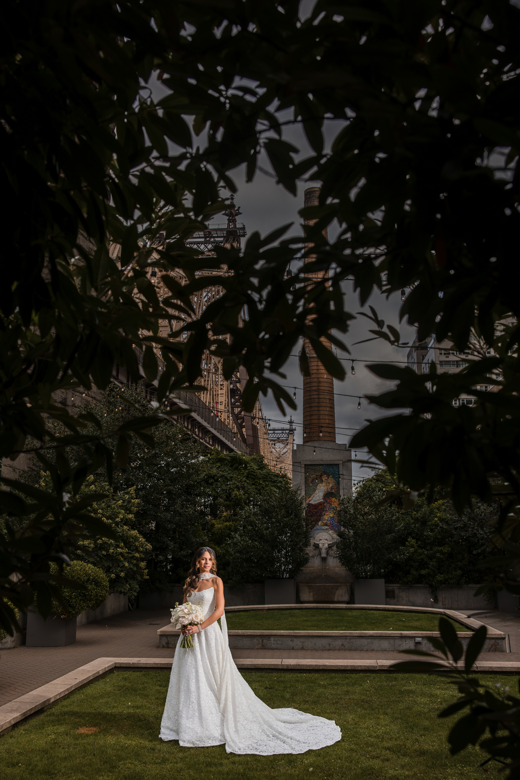 Bride in a white gown stands on a lawn holding a bouquet, framed by tree branches, with urban buildings and a mural in the background, capturing the elegant charm of a Guastavino’s wedding.