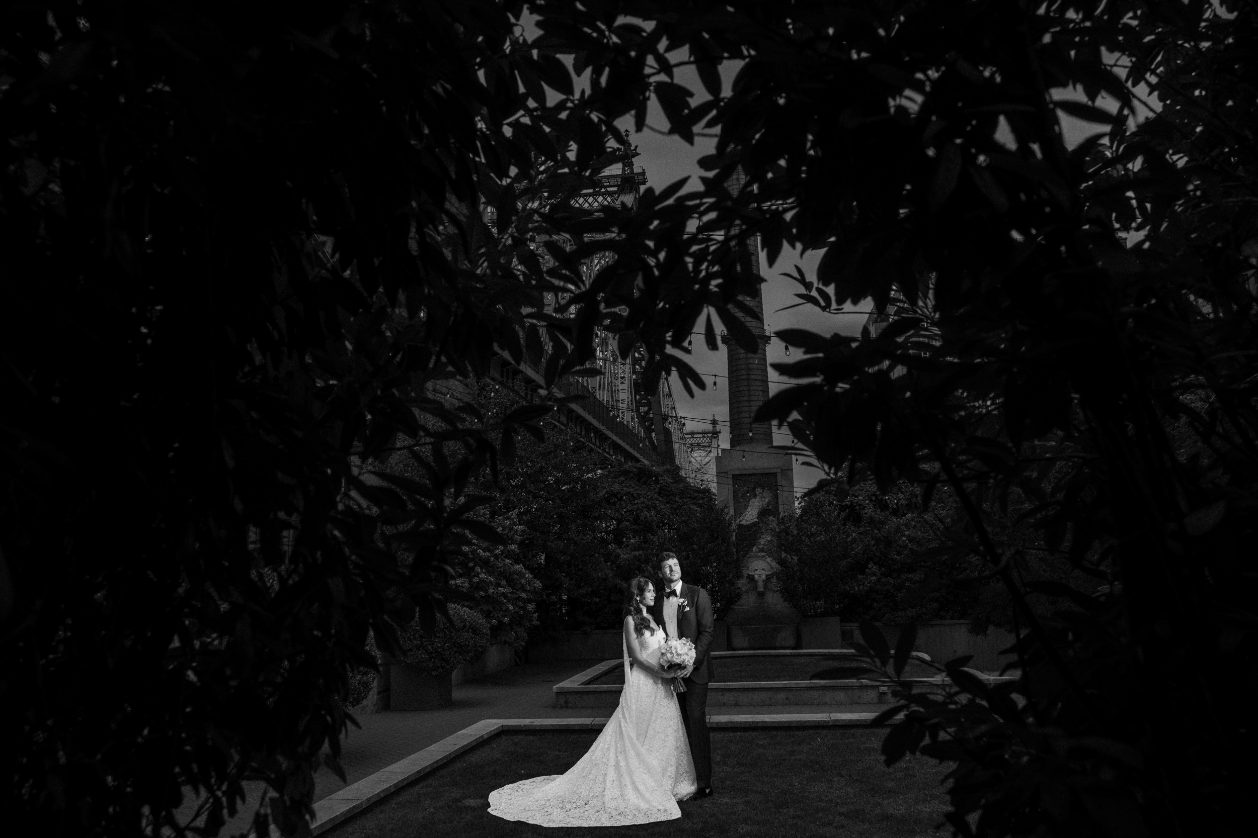 A bride and groom stand together in a garden, framed by dark foliage, with city buildings visible in the background, capturing the romantic ambiance of a Guastavino’s wedding.