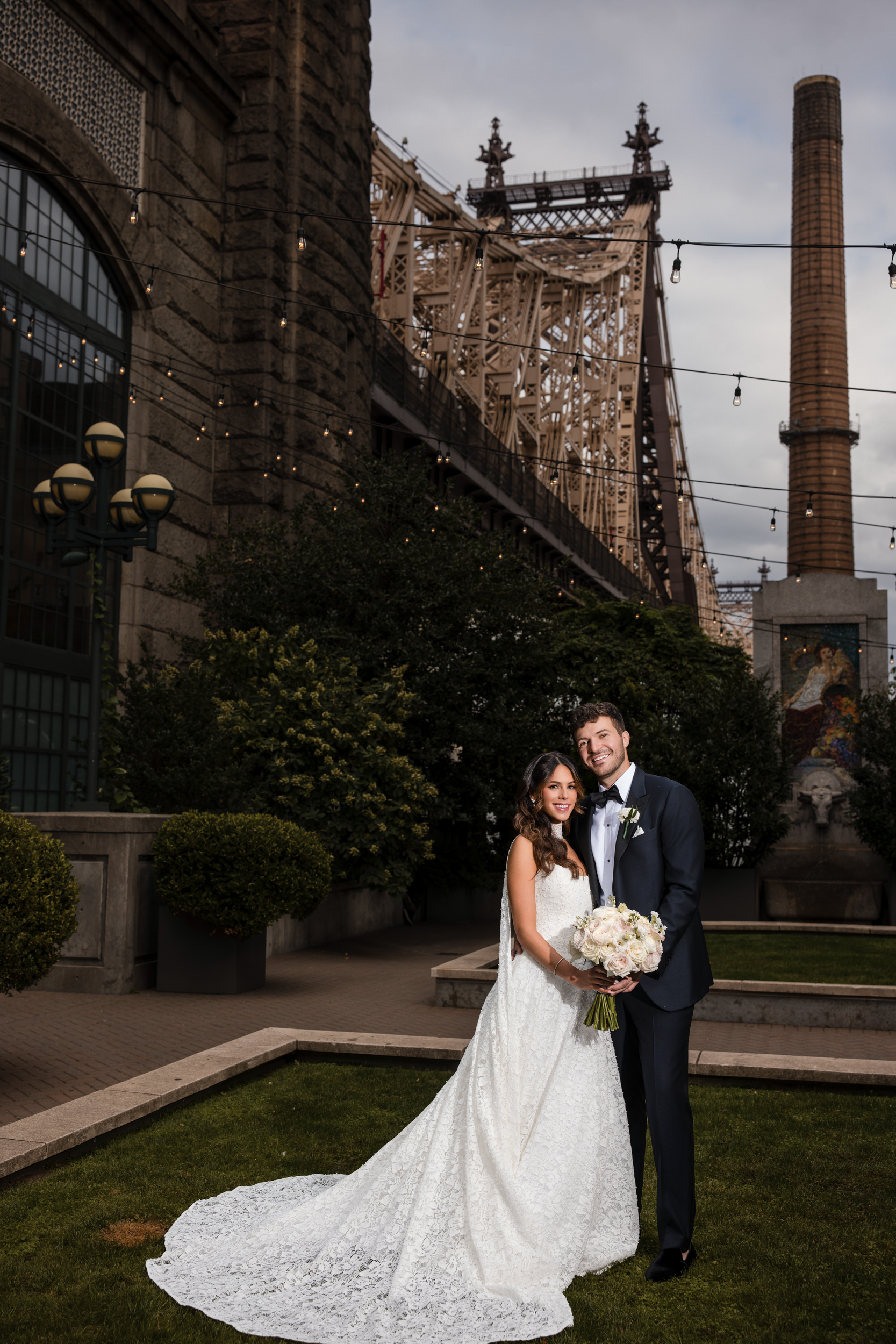 A bride and groom stand together outdoors, smiling and holding a bouquet, with a large bridge and industrial structures in the background, evoking the timeless elegance of a Guastavino’s wedding.
