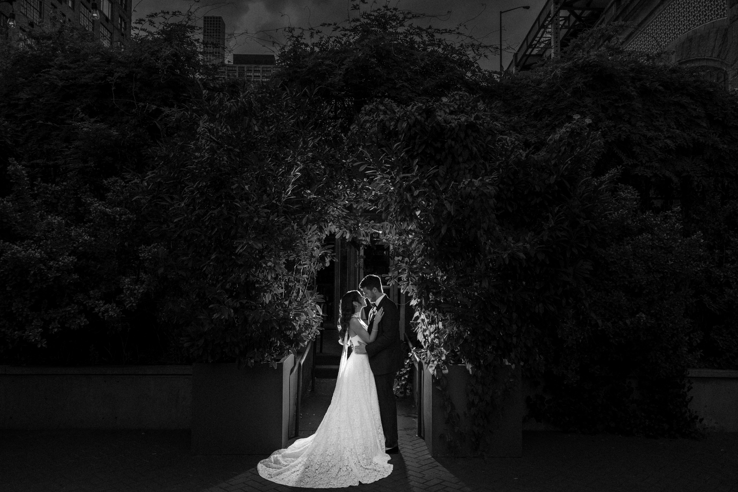 A bride and groom stand close together under a leafy archway, illuminated by sunlight, surrounded by shadows in an outdoor setting, capturing the timeless romance of a Guastavino’s wedding.