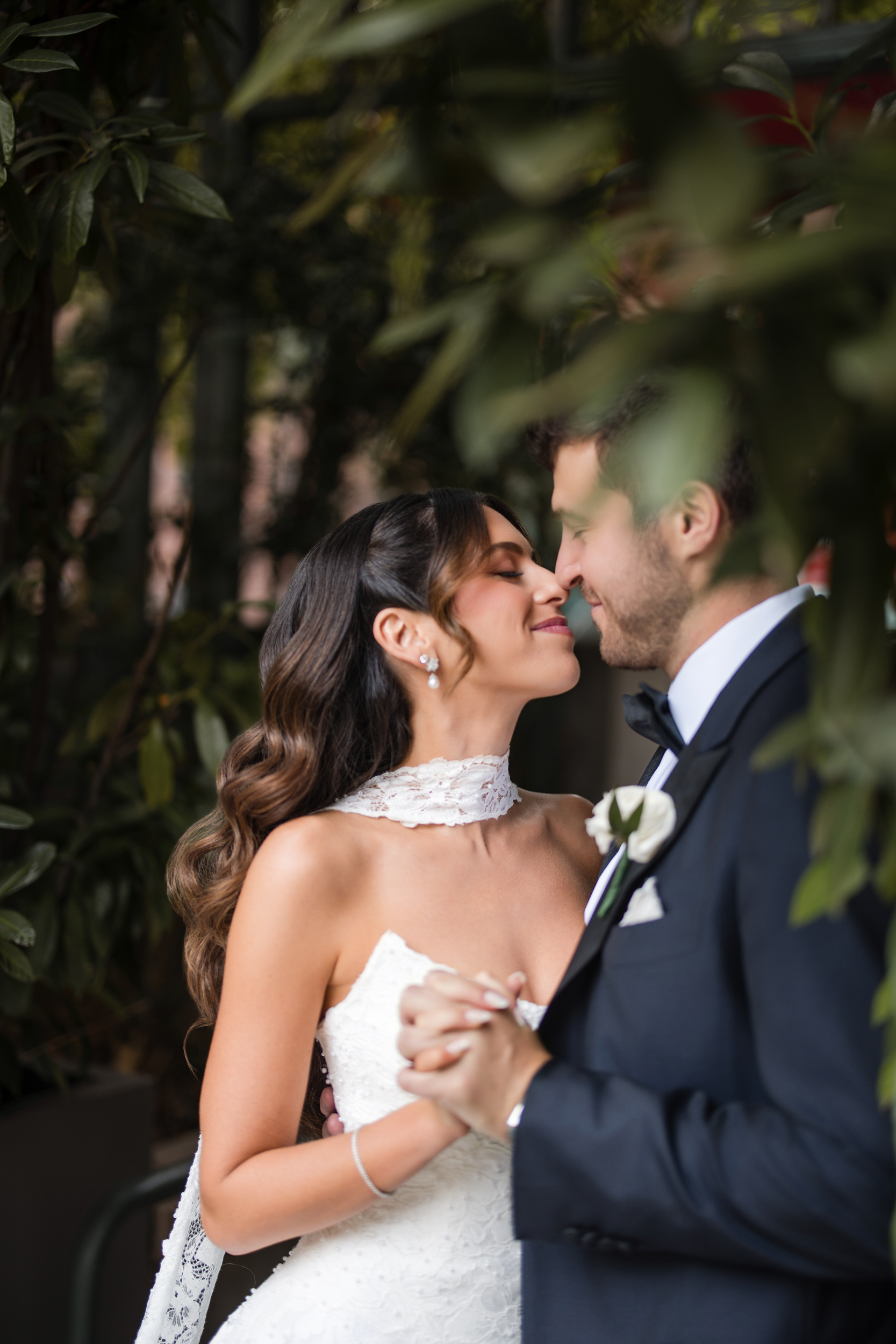 A bride and groom stand close together at their Guastavino’s wedding, holding hands and smiling with their noses almost touching, surrounded by greenery.