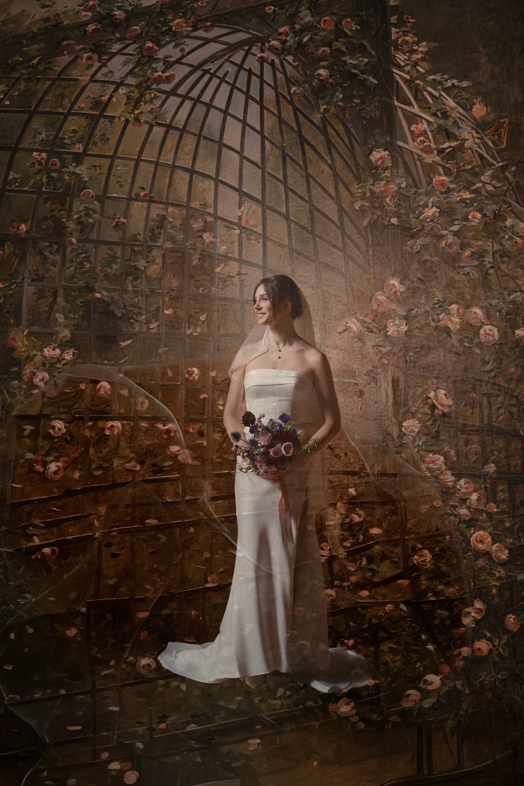 A Lotte New York Palace wedding bride in a white dress holds a bouquet as she stands in front of a backdrop adorned with roses and a metal garden trellis.