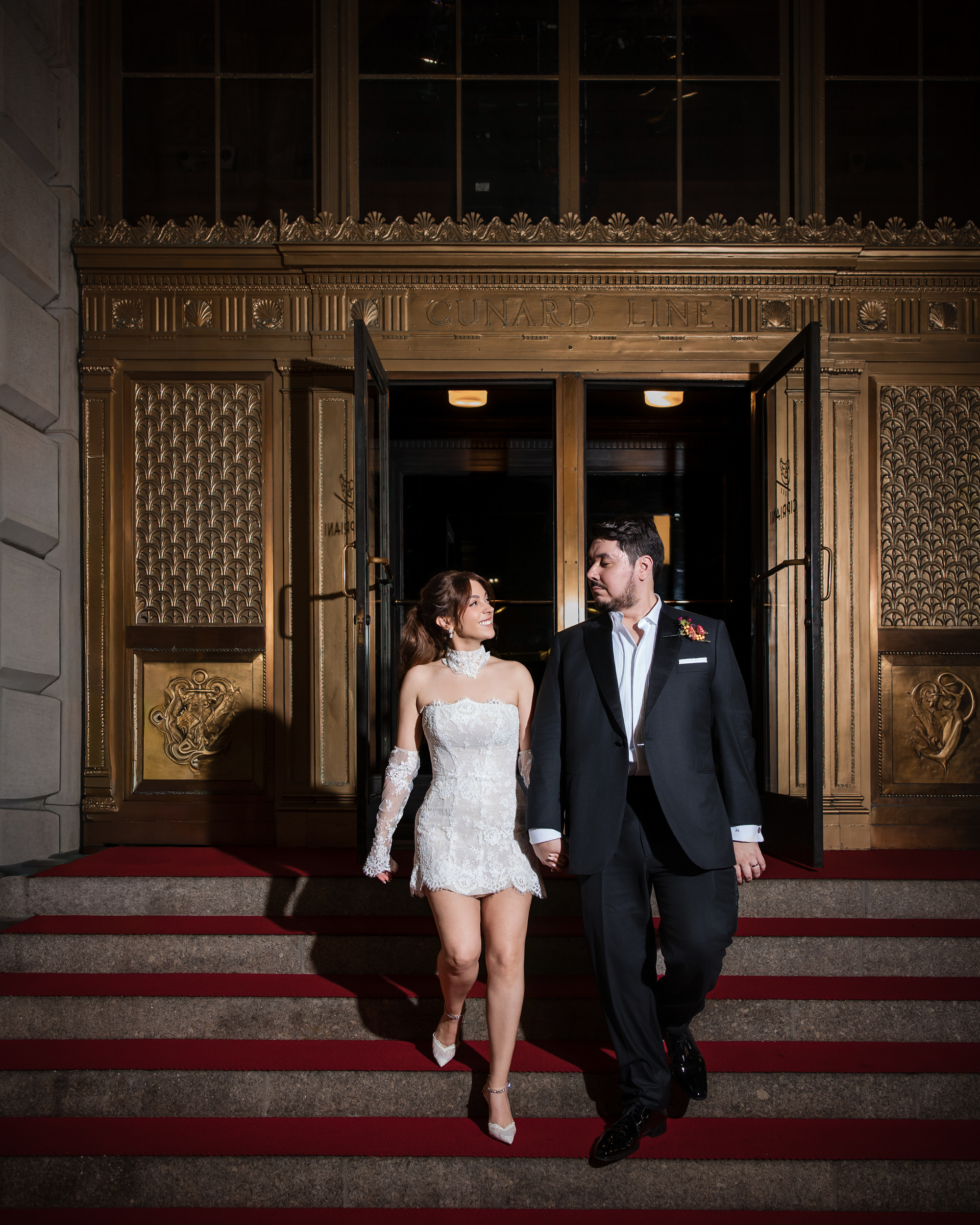A couple in wedding attire walks down the red carpeted steps in front of the Cipriani 25 Broadway entrance.