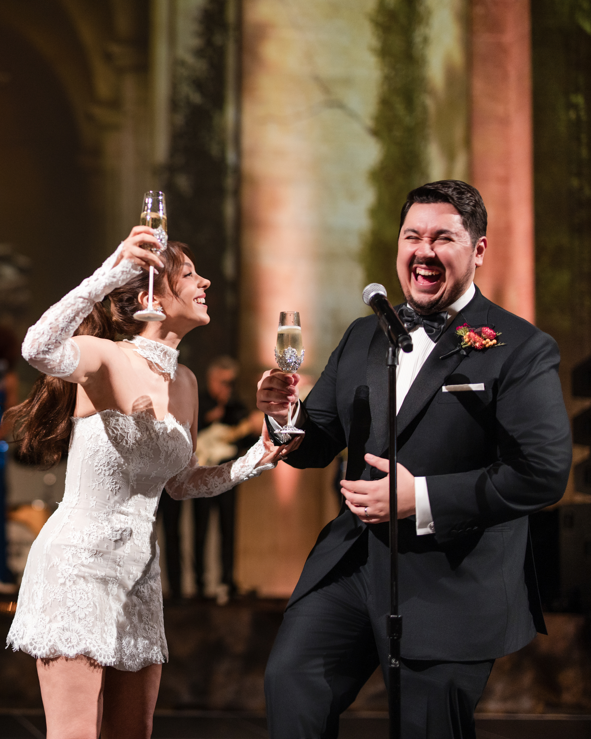 A bride and groom laughing and holding up champagne glasses at a wedding reception.