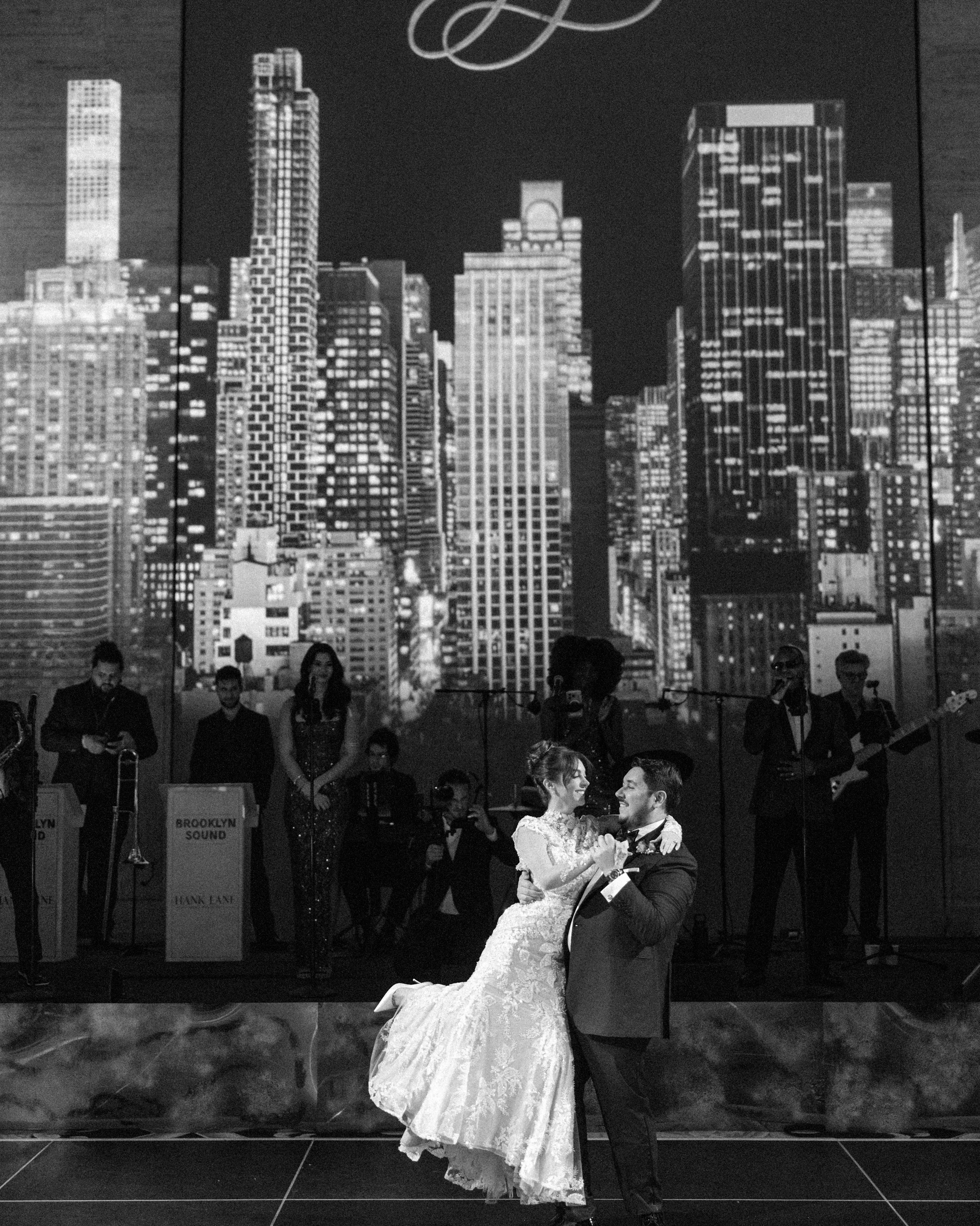 A bride and groom dance on stage at a Cipriani 25 Broadway wedding, set before a live band and a large backdrop of a city skyline.