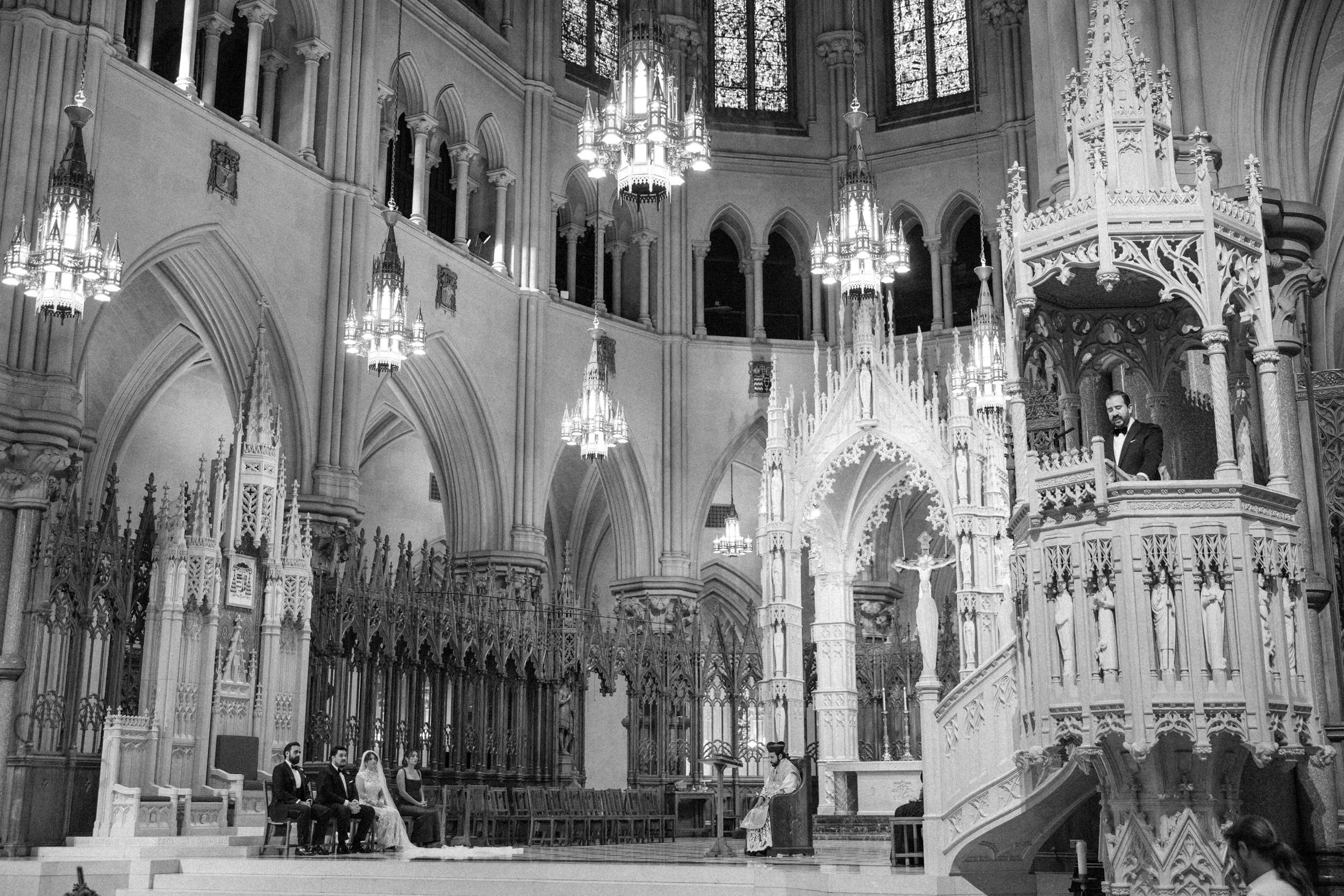 A wedding ceremony at Cipriani 25 Broadway, with a couple seated at the altar as an officiant speaks from an elevated stone pulpit inside the cathedral. A wedding ceremony at Cipriani 25 Broadway, with a couple seated at the altar as an officiant speaks from an elevated stone pulpit inside the cathedral.