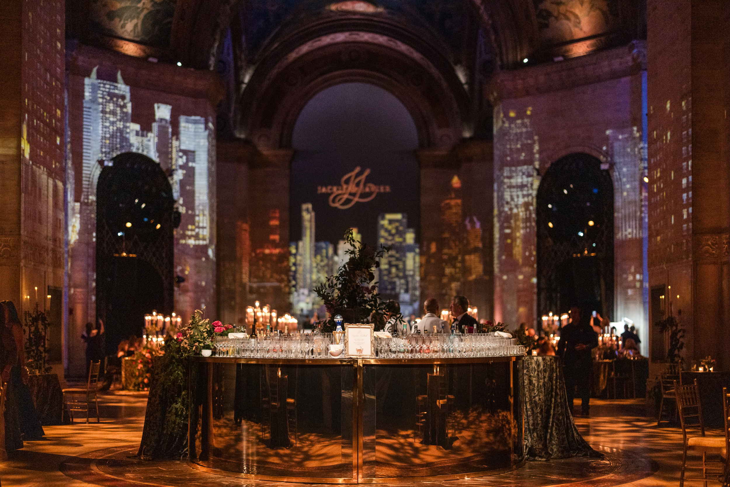A circular bar in a dimly lit Cipriani 25 Broadway hall, featuring a cityscape projection on the walls and an illuminated monogram backdrop. A circular bar in a dimly lit Cipriani 25 Broadway hall, featuring a cityscape projection on the walls and an illuminated monogram backdrop.