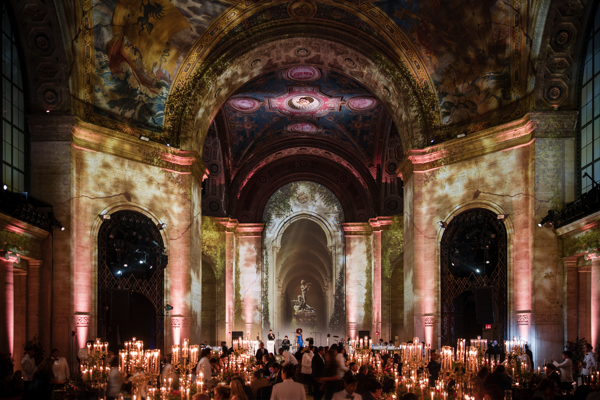 A grand hall at Cipriani 25 Broadway with vaulted, muraled ceilings and stone arches, set up for a dinner event with numerous tall candles on tables. A grand hall at Cipriani 25 Broadway with vaulted, muraled ceilings and stone arches, set up for a dinner event with numerous tall candles on tables.