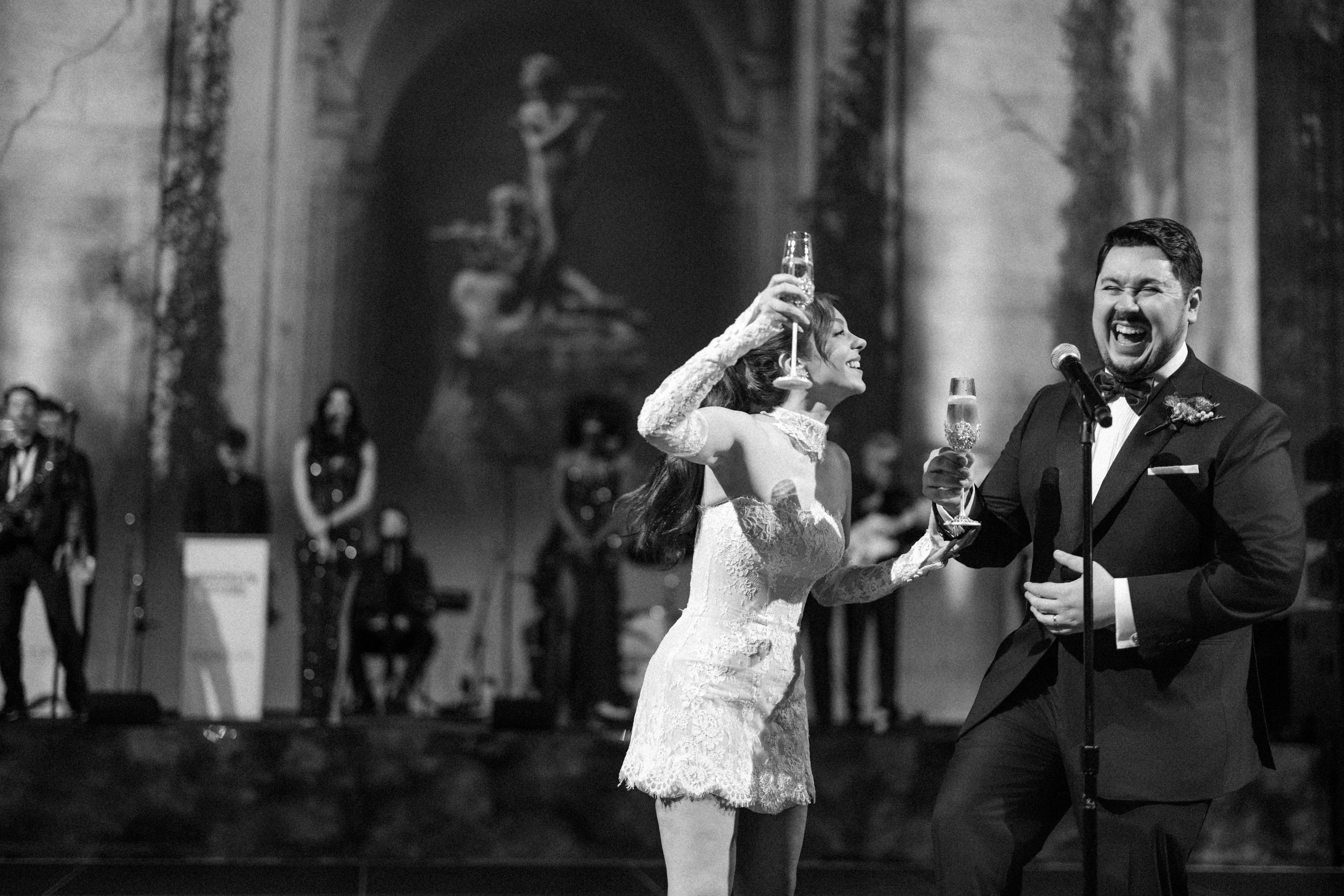 A bride and groom laughing and raising champagne flutes on stage at a Cipriani 25 Broadway wedding. A bride and groom laughing and raising champagne flutes on stage at a Cipriani 25 Broadway wedding.