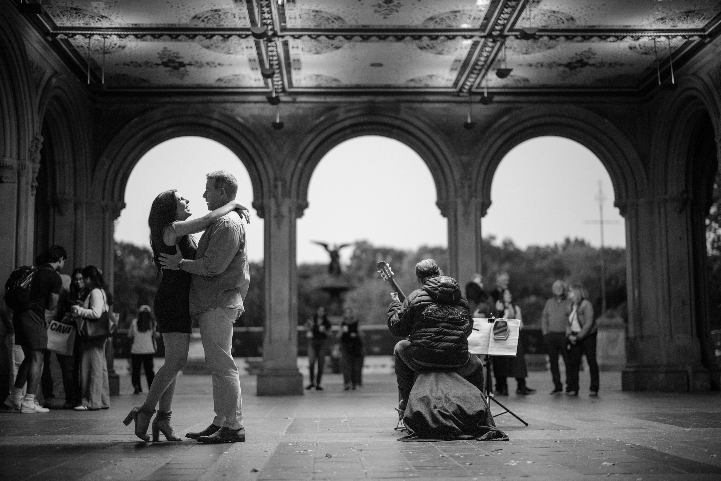 A couple dances in Bethesda Terrace as a street musician plays guitar during a Central Park proposal.