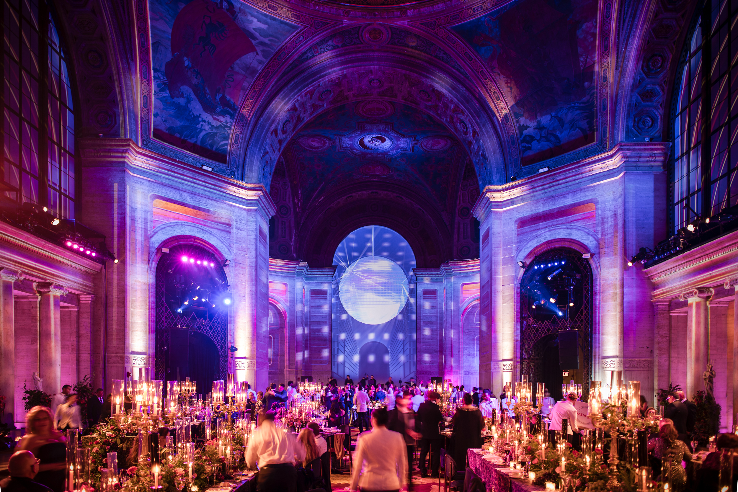A Cipriani 25 Broadway wedding gala dinner inside a grand hall with arched ceilings, murals, tables with tall candles, and a large disco ball suspended in the center. A Cipriani 25 Broadway wedding gala dinner inside a grand hall with arched ceilings, murals, tables with tall candles, and a large disco ball suspended in the center.