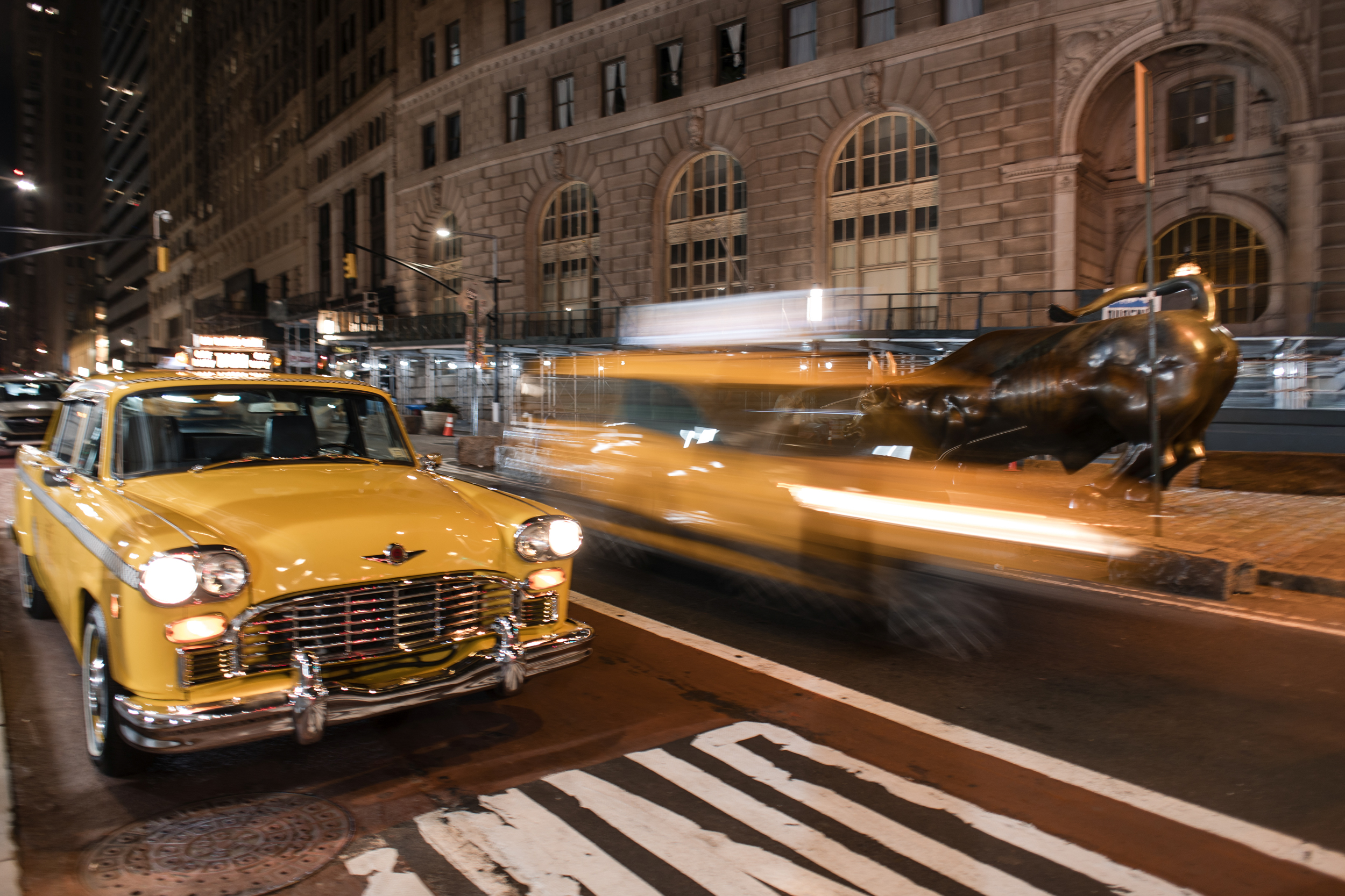 A vintage yellow taxi parked on a city street near the Charging Bull statue by Cipriani 25 Broadway, with a motion-blurred taxi driving past in the background. A vintage yellow taxi parked on a city street near the Charging Bull statue by Cipriani 25 Broadway, with a motion-blurred taxi driving past in the background.