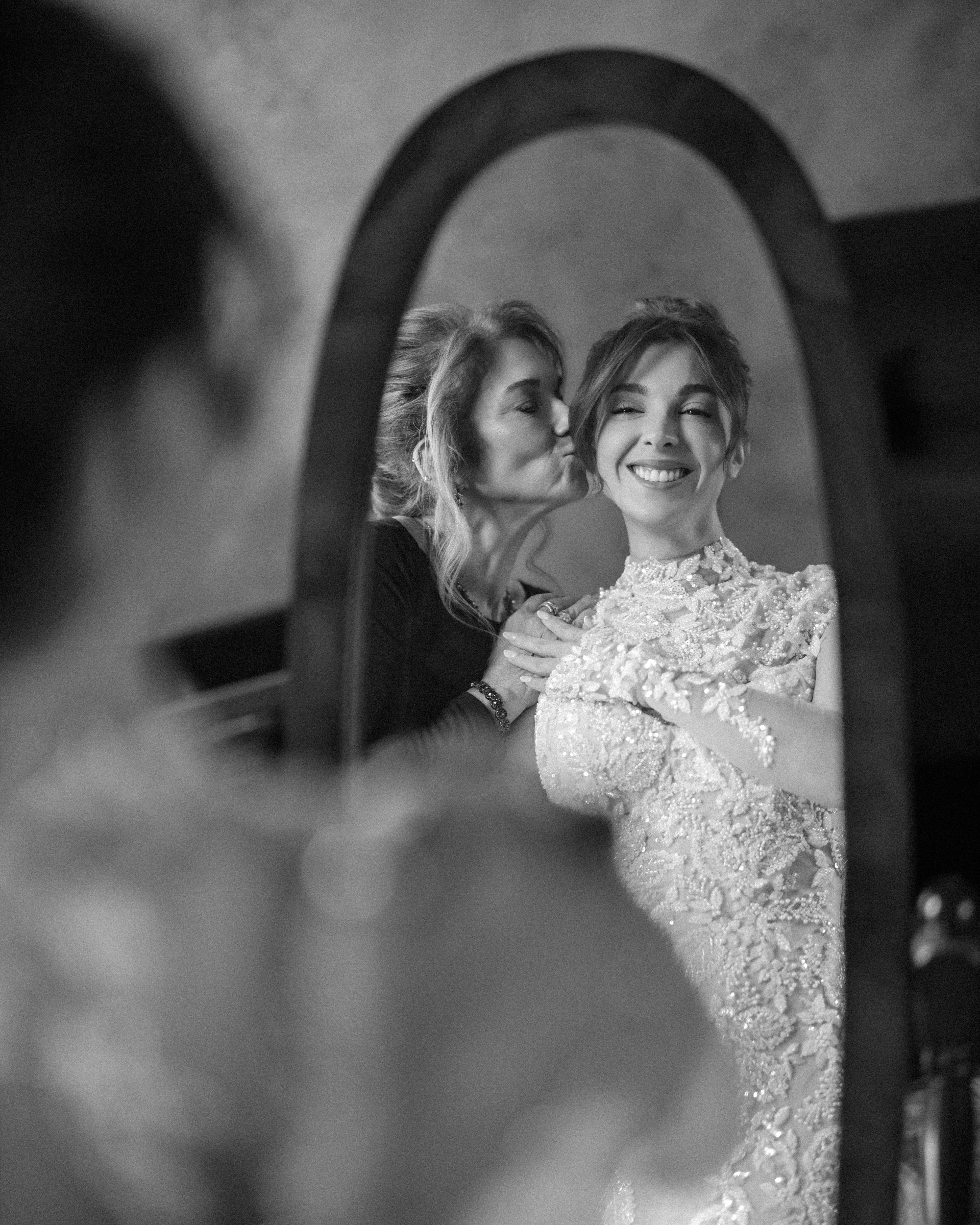 A smiling bride in her gown looks into a mirror at Cipriani 25 Broadway as another woman kisses her on the cheek.
