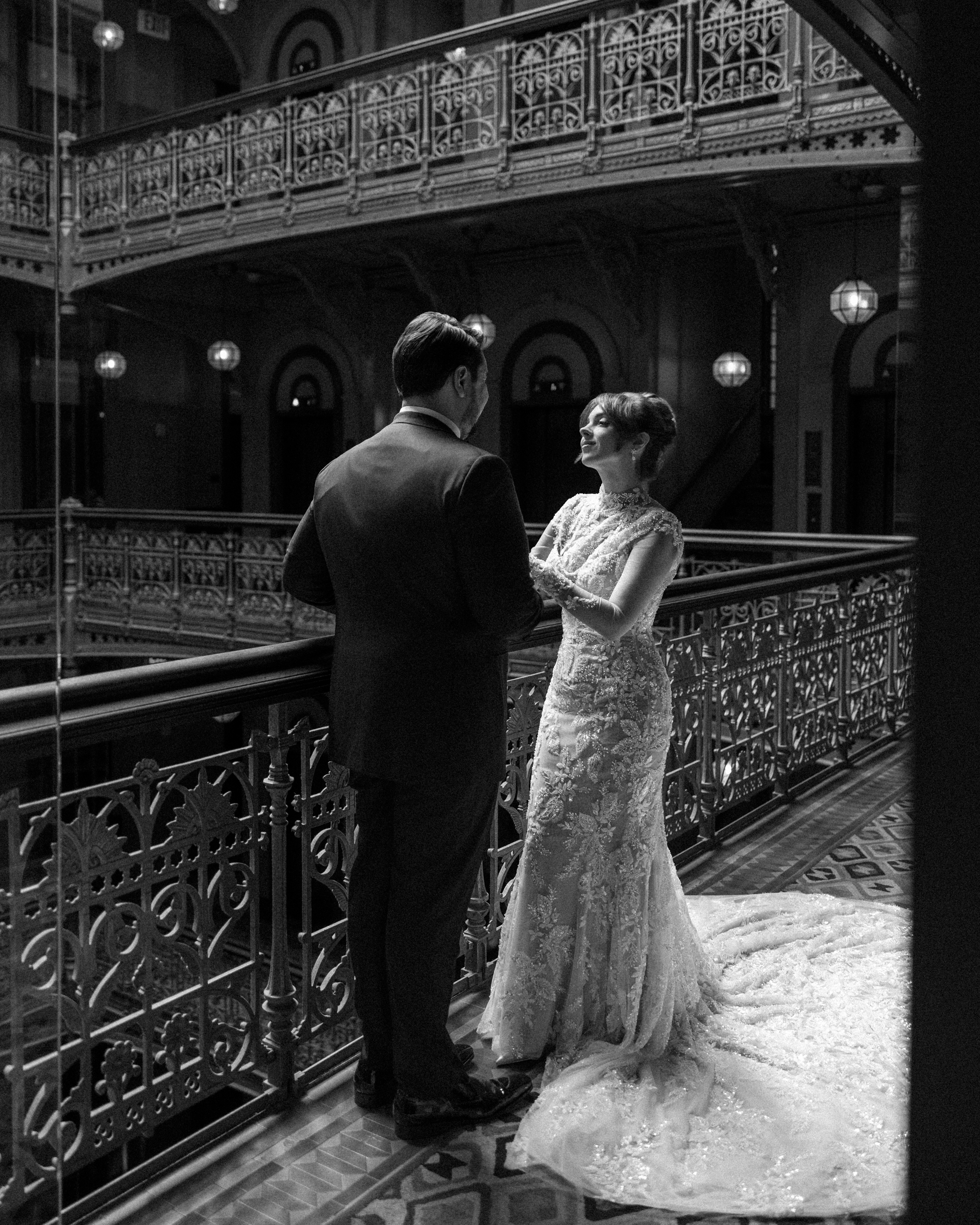 A bride and groom face each other on a balcony with ornate metal railings inside a historic Cipriani 25 Broadway building.